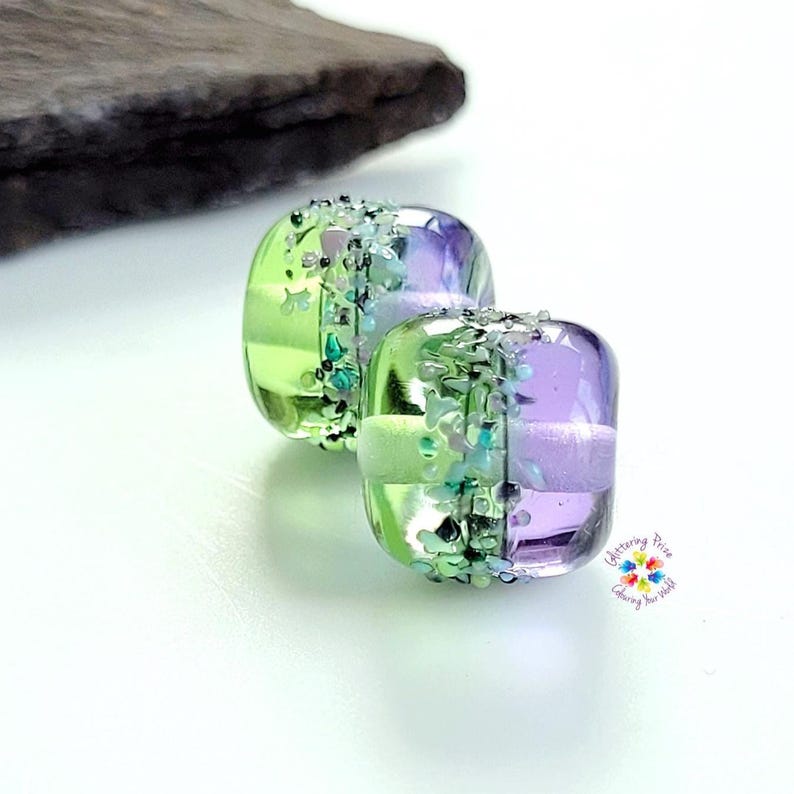 May include: Two square glass beads featuring a split color design of lime green and lavender purple. A central band of textured, multi-colored crushed glass fragments separates the two colors. The logo Glittering Prize Colouring Your World is visible.