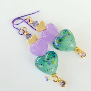 Puede incluir: Pendientes de cristal en forma de corazón de color morado y verde con detalles dorados y cuentas de cristal morado.