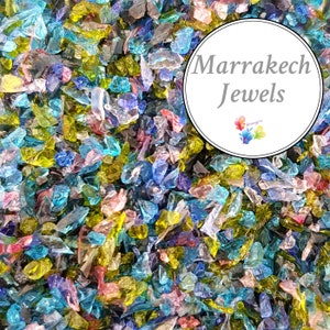 May include: A close-up of a pile of colorful, iridescent glass pieces. The glass pieces are various shapes and sizes, and they are all very sparkly. The text "Marrakech Jewels" is written in a white circle on top of the glass pieces.