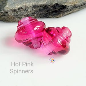 Könnte beinhalten: Zwei pinkfarbene Spinner mit einem Wirbeldesign. Die Spinner bestehen aus einem durchscheinenden Material und sind vor einem weißen Hintergrund platziert. Der Text "Hot Pink Spinners" ist sichtbar.