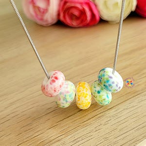 Peut inclure: Un collier à chaîne argentée avec six perles colorées et mouchetées. Les perles sont roses, jaunes, vertes et bleues. Le collier est présenté sur une surface en bois, avec des roses roses et blanches floues en arrière-plan.