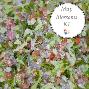 Op de afbeelding: Een close-up van een verscheidenheid aan kleine, kleurrijke glazen stukjes. De stukjes zijn in tinten groen, roze en helder. De tekst "May Blossoms K1" is zichtbaar in de afbeelding.