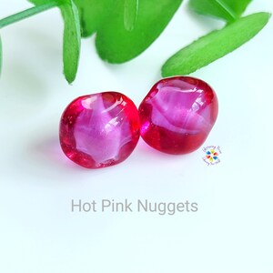May include: Two hot pink, rounded glass nuggets with a translucent appearance. The nuggets are a vibrant pink colour and have a smooth, polished surface. The text "Hot Pink Nuggets" is displayed below the nuggets.