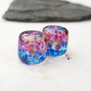 May include: Two colourful glass beads with a cylindrical shape. The beads feature a blend of blue, purple, pink, and yellow hues, with a textured, speckled appearance. The beads are set against a white background.