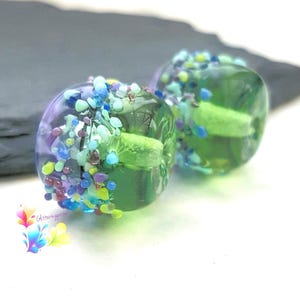 May include: Two round glass beads featuring a translucent green and purple base, decorated with a textured band of small, multi-colored glass fragments in shades of blue, yellow, and purple, with a central light green core and a logo.