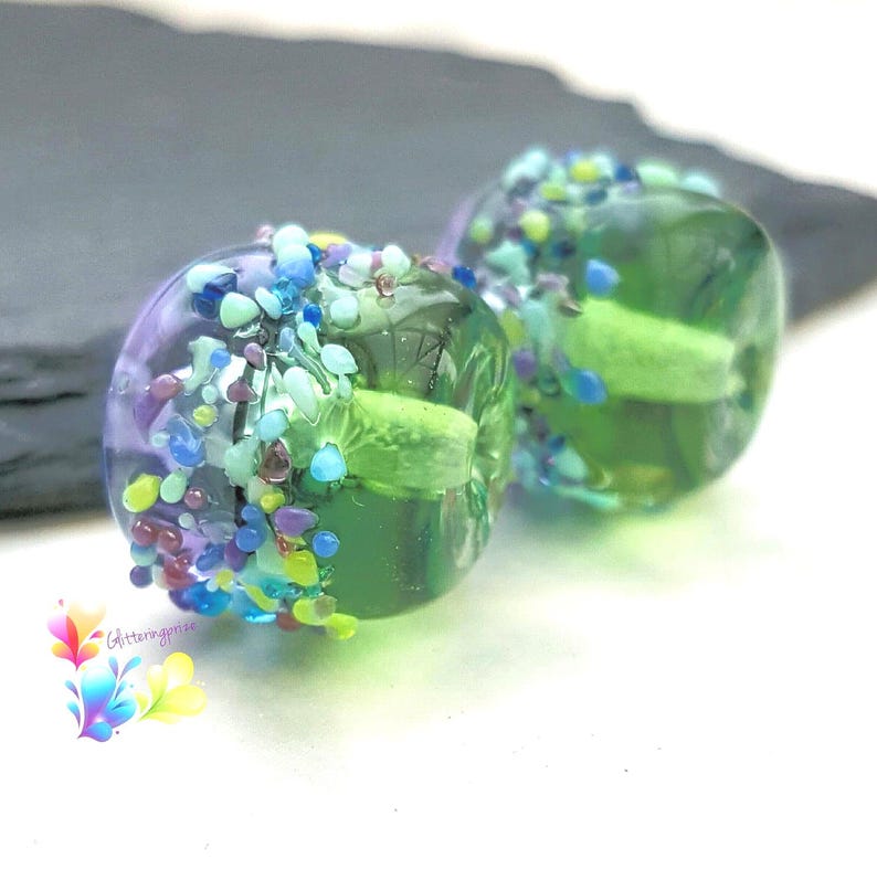 May include: Two round glass beads featuring a translucent green and purple base, decorated with a textured band of small, multi-colored glass fragments in shades of blue, yellow, and purple, with a central light green core and a logo.