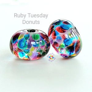 May include: Two colourful glass beads with the text "Ruby Tuesday Donuts". The beads are a mix of red, blue, green, and purple, with a clear background. One bead has a hole through the centre.