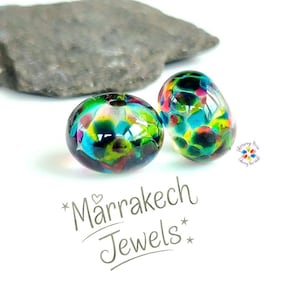 May include: Two round glass beads featuring a multi-colored pattern of blue, green, yellow, and red spots. The text "Mårrakech Jewels" is printed below the beads, along with a small logo featuring a colorful flower design and text.