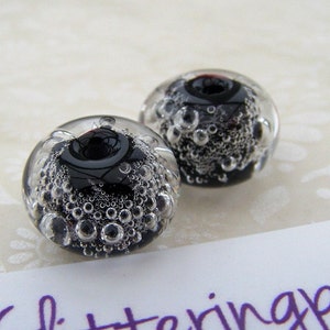 Puede incluir: Dos cuentas de vidrio transparente con un centro oscuro y brillante y numerosas burbujas diminutas suspendidas en su interior. Las cuentas son redondas y parecen hechas a mano, con un diseño artístico único. Fondo claro y estampado.