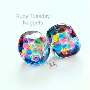 Può includere: Due pepite di vetro colorate con il testo "Ruby Tuesday Nuggets". Le pepite sono un mix di colori vivaci tra cui blu, rosa, verde e arancione. L'immagine include anche un logo con il testo "Glittering Prize Colouring Your World".