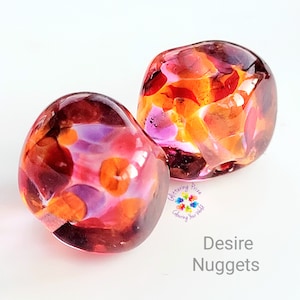 Peut inclure: Deux pépites de verre aux couleurs vives, notamment orange, violet et rouge. Les pépites arrondies ont une finition brillante. Le texte "Desire Nuggets" est visible. Idéal pour la décoration.