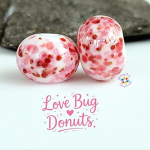 Könnte beinhalten: Zwei runde, donut-förmige Glasperlen mit weißer Basis und einem gesprenkelten Muster aus rosa und roten Flecken. Der Text "Love Bug Donuts" ist in Rosa unter den Perlen aufgedruckt, zusammen mit einem kleinen Logo mit bunten Blumenformen.