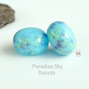 May include: Two blue glass donut beads with a speckled pattern of green and purple. The beads are smooth and glossy, with the text "Paradise Sky Donuts" below them.