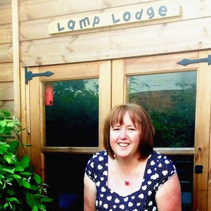 May include: A woman smiles in front of a wooden structure with the words "Lomp Lodge" on a sign above the door. She wears a blue and white polka dot dress and a red pendant necklace. The background includes greenery.