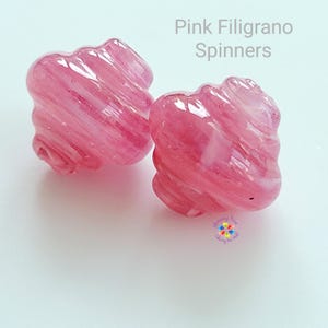 Könnte beinhalten: Zwei rosa Filigrano-Spinner mit einem Wirbeldesign. Die Spinner haben eine leuchtend rosa Farbe und eine glänzende Oberfläche. Der Text "Pink Filigrano Spinners" wird oben im Bild angezeigt.