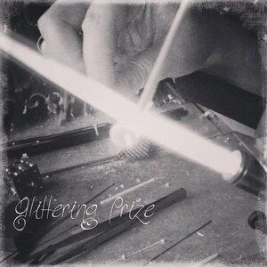 May include: A black and white image showing a glassblowing process with a glowing glass rod being shaped. The text "Glittering Prize" is written in a cursive script across the bottom. Various metal tools and thin glass rods are visible.