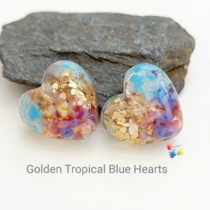 Pode incluir: Dois enfeites de vidro em forma de coração com tema tropical. Os enfeites apresentam uma mistura de cores azul, dourado, vermelho e roxo. O texto "Golden Tropical Blue Hearts" é exibido abaixo.