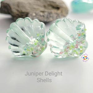 May include: Two light blue glass seashells with iridescent glitter and a white background. The text "Juniper Delight Shells" is below the shells.