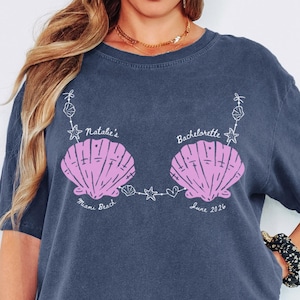 Puede incluir: Camiseta azul marino con diseño de conchas rosas. El texto en la camiseta dice "Natalie's Bachelorette" con "Miami Beach" y "June 2026" debajo. La camiseta es de corte holgado.