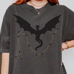 May include: A dark grey t-shirt featuring a black silhouette of a dragon and a constellation of stars in gold.