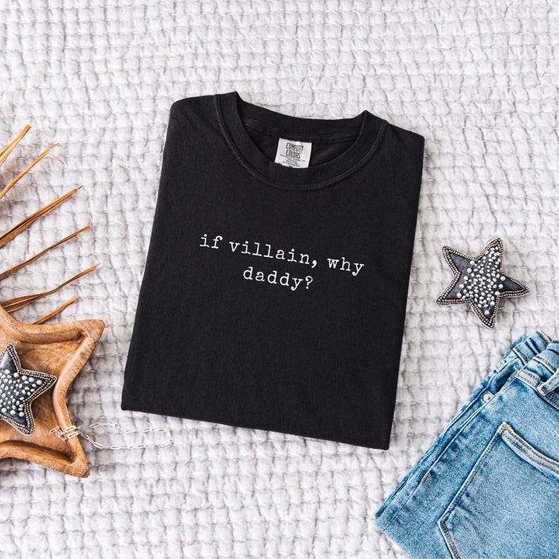 May include: Black t-shirt with the text "if villain, why daddy?" in white. The shirt is displayed on a white textured surface, next to a pair of blue jeans and a star-shaped decorative item.