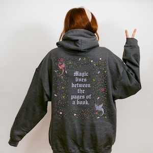 May include: Dark gray hoodie with the text "Magic lives between the pages of a book." The back of the hoodie features a floral design with pink and yellow accents, and two mythical creatures. The hoodie has a hood and long sleeves.