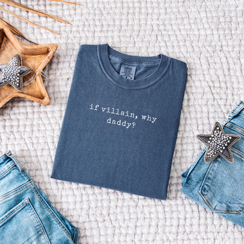 May include: A slate blue t-shirt with the phrase "if villain, why daddy?" in white lettering. The shirt is folded and placed on a textured surface, next to a pair of blue jeans and star-shaped decorations.