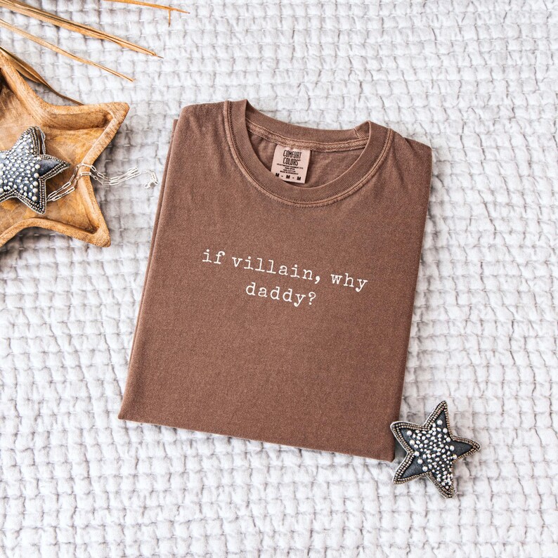 May include: A brown t-shirt with the text "if villain, why daddy?" in white. The shirt is folded and lies on a white, textured surface. A wooden star-shaped dish and a star-shaped ornament are in the background.