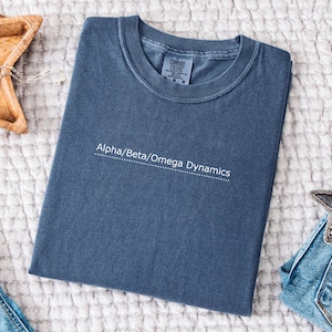 May include: A folded, blue Comfort Colors t-shirt with the text "Alpha/Beta/Omega Dynamics" in white. The shirt is on a white, textured surface.