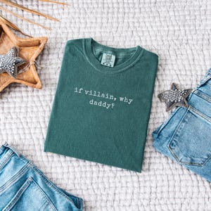 May include: A teal-colored t-shirt with the phrase "if villain, why daddy?" in white lettering. The shirt is folded and placed on a textured white surface, with a pair of blue jeans and decorative stars nearby.