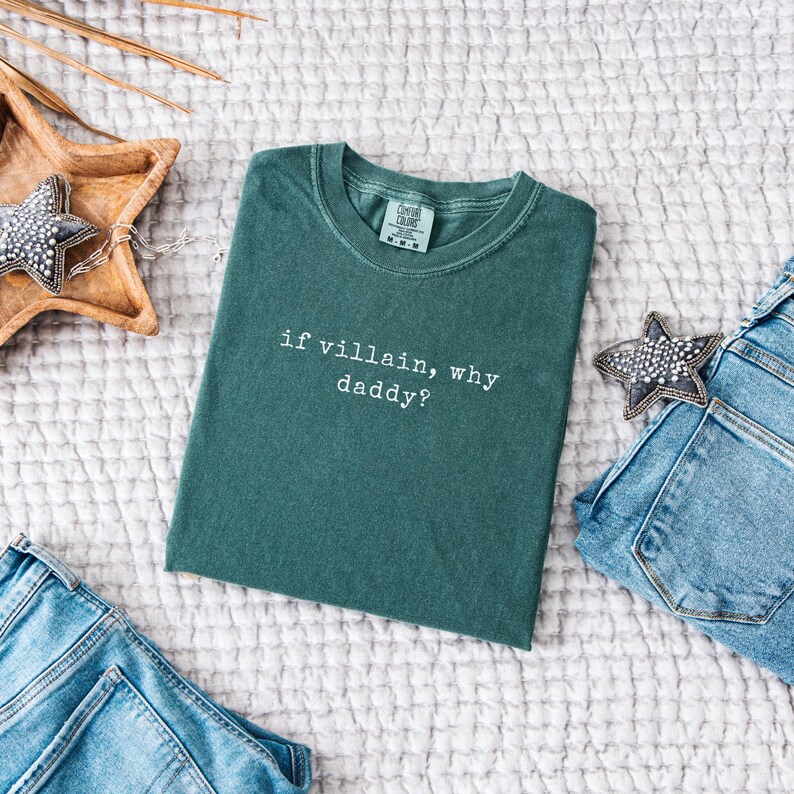 May include: A teal-colored t-shirt with the phrase "if villain, why daddy?" in white lettering. The shirt is folded and placed on a textured white surface, with a pair of blue jeans and decorative stars nearby.