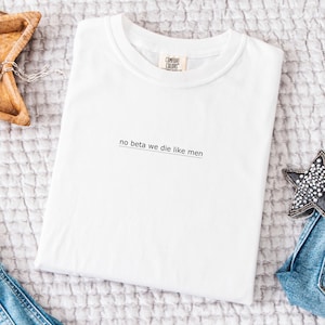 May include: White Comfort Colors t-shirt with the text "no beta we die like men" in a simple, black font. The shirt is folded neatly on a textured, light-colored surface. A small tag is visible near the collar.