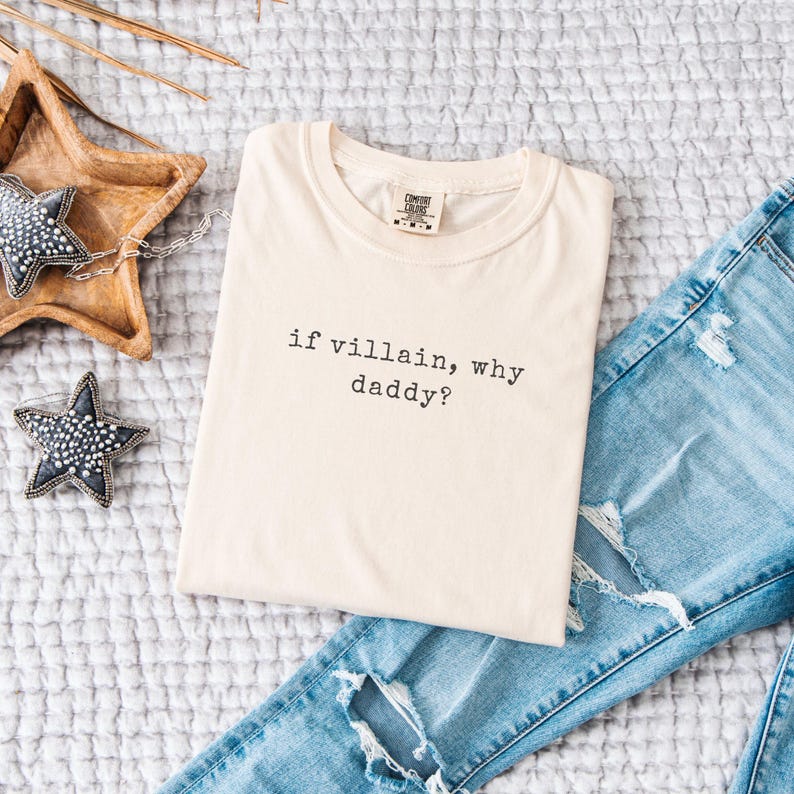 May include: Cream-colored t-shirt with the phrase "if villain, why daddy?" in black lettering. The shirt is folded and placed on a textured, light gray surface next to a pair of blue jeans with distressed details. Decorative star-shaped ornaments are also present.