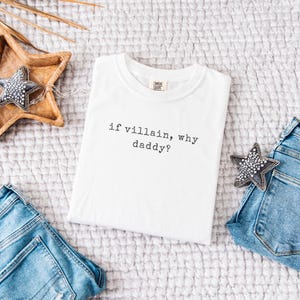 May include: White t-shirt with the text "if villain, why daddy?" in black. The shirt is displayed on a textured surface, with a wooden star-shaped tray and denim jeans visible. A decorative star is also present.