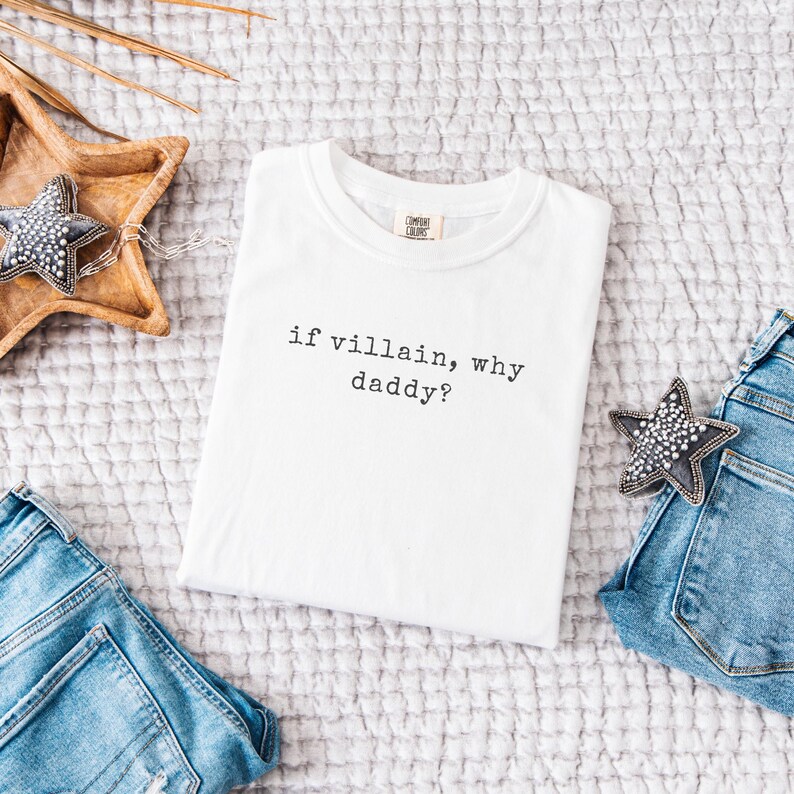 May include: White t-shirt with the text "if villain, why daddy?" in black. The shirt is displayed on a textured surface, with a wooden star-shaped tray and denim jeans visible. A decorative star is also present.