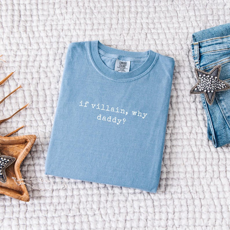 May include: A light blue t-shirt with the text "if villain, why daddy?" in white. The shirt is folded on a white textured surface, next to a pair of blue jeans and a star-shaped accessory.