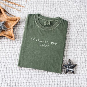 May include: An olive green t-shirt with the phrase "if villain, why daddy?" in white text. The shirt is folded and lies on a textured white surface. A decorative star and wooden bowl are also visible.
