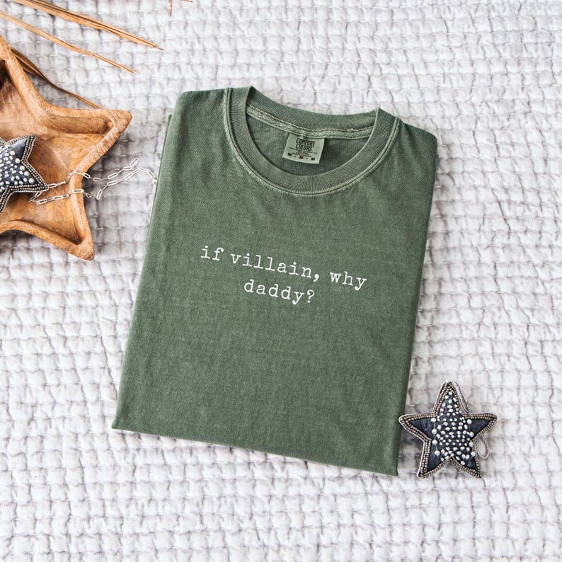 May include: An olive green t-shirt with the phrase "if villain, why daddy?" in white text. The shirt is folded and lies on a textured white surface. A decorative star and wooden bowl are also visible.