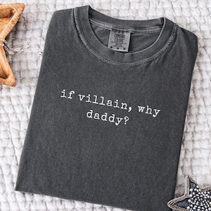 May include: Dark grey t-shirt with the text "if villain, why daddy?" in white. The shirt has a crew neck and a tag from Comfort Colors. The shirt is folded on a white, textured surface.