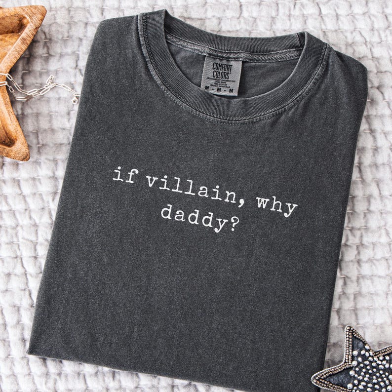 May include: Dark grey t-shirt with the text "if villain, why daddy?" in white. The shirt has a crew neck and a tag from Comfort Colors. The shirt is folded on a white, textured surface.