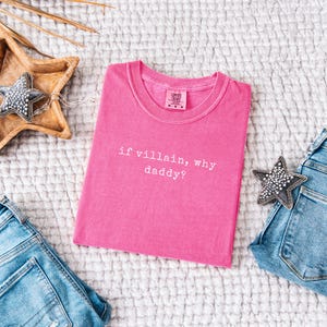 May include: A bright pink t-shirt with the phrase "if villain, why daddy?" in white text. The shirt is laid flat on a textured white surface, with a pair of blue jeans and decorative stars nearby. The shirt is a Comfort Colors brand.