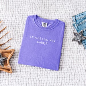 May include: A lavender Comfort Colors t-shirt with the phrase "if villain, why daddy?" in white text. The shirt is laid flat on a textured white surface, with a pair of blue jeans and a star-shaped decorative item.