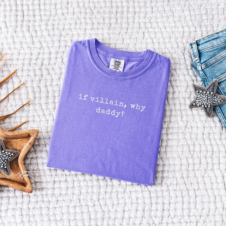 May include: A lavender Comfort Colors t-shirt with the phrase "if villain, why daddy?" in white text. The shirt is laid flat on a textured white surface, with a pair of blue jeans and a star-shaped decorative item.