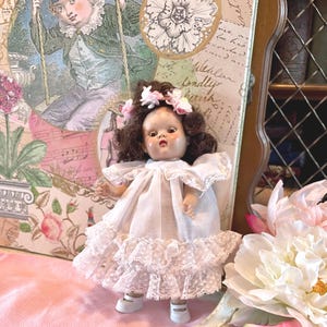 May include: A vintage doll with brown hair and a floral crown, wearing a white dress with lace trim. The doll is posed in front of a decorative background with a floral and vintage illustration theme. The doll is wearing white shoes.