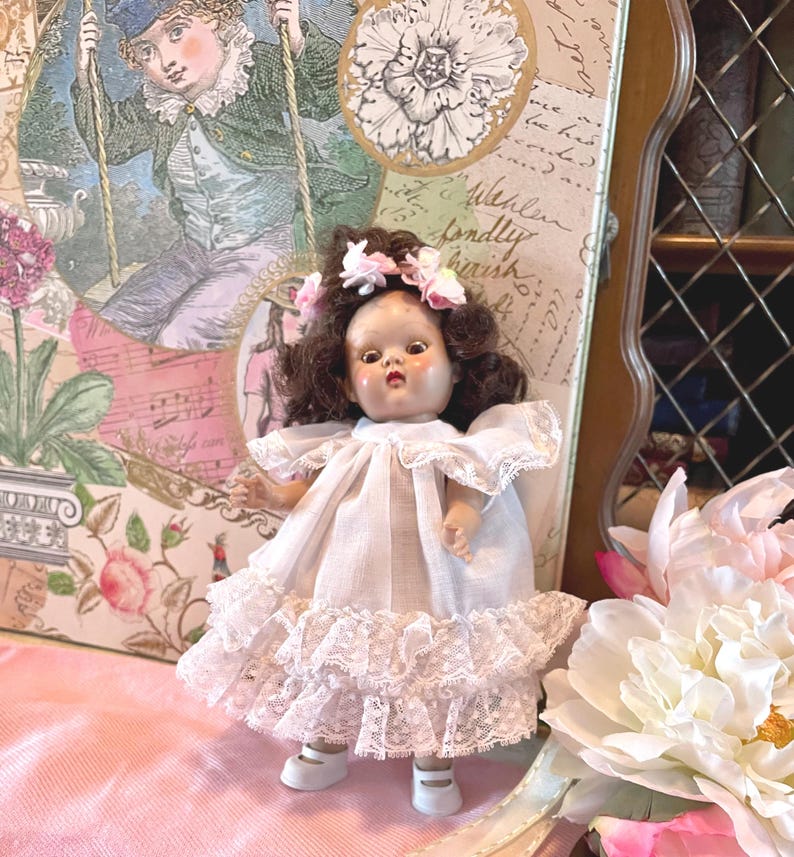 May include: A vintage doll with brown hair and a floral crown, wearing a white dress with lace trim. The doll is posed in front of a decorative background with a floral and vintage illustration theme. The doll is wearing white shoes.