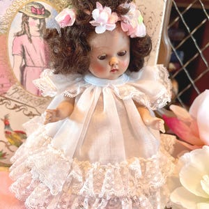 May include: A vintage doll with brown hair and a floral crown, wearing a white dress with lace trim. The doll has rosy cheeks and is posed in front of a vintage illustration.