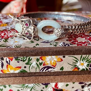 May include: Silver-toned bracelet with a dragon head design, featuring a light blue oval stone and small red stones. The bracelet has a chain-like texture and is displayed on a floral patterned surface.