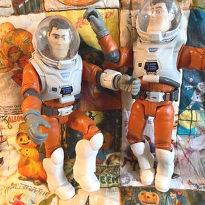 May include: Two Buzz Lightyear action figures in orange and white space suits with clear helmets. Each figure has the number "15" on the chest. The figures are posed on a Halloween-themed quilt with pumpkin and witch illustrations.