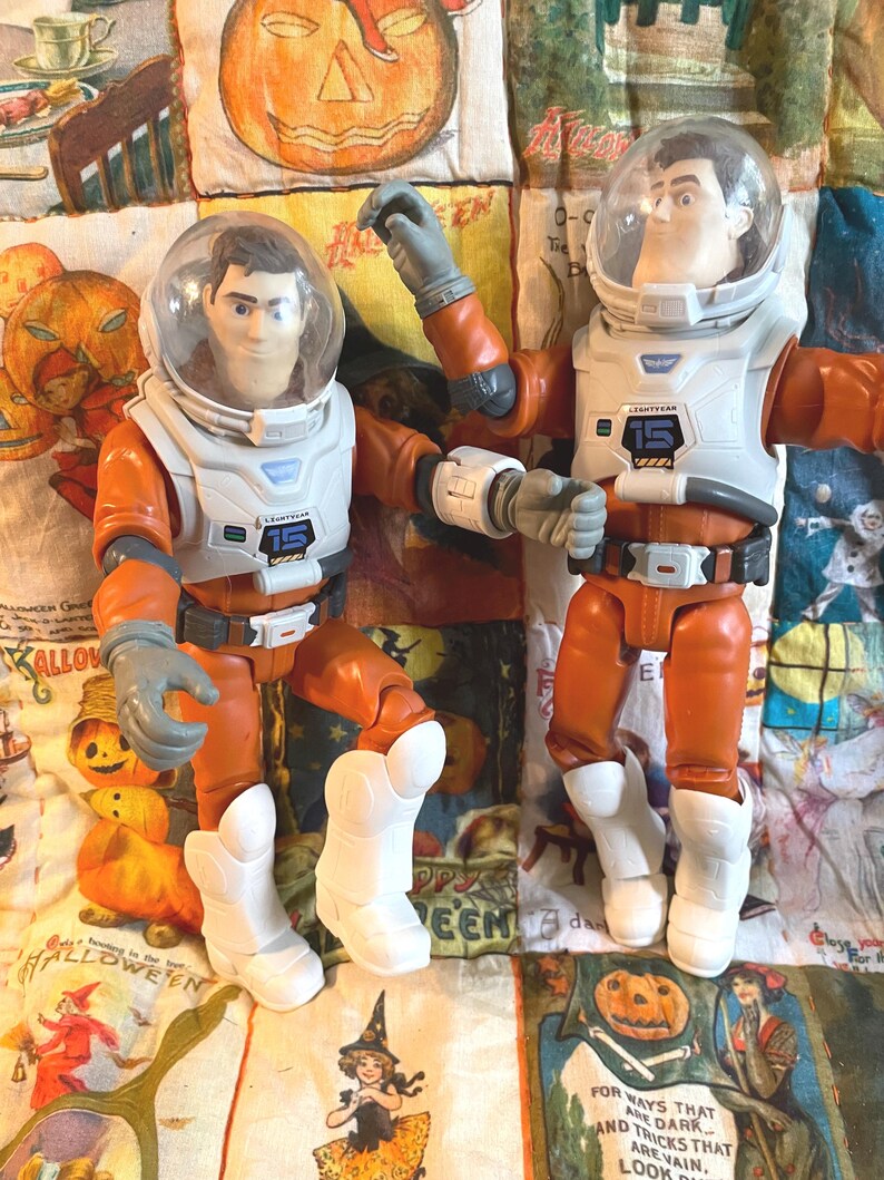 May include: Two Buzz Lightyear action figures in orange and white space suits with clear helmets. Each figure has the number "15" on the chest. The figures are posed on a Halloween-themed quilt with pumpkin and witch illustrations.