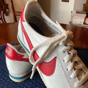 May include: White leather Nike trainers with a red swoosh and blue trim. The shoes have white laces and the text "SIZE 5 81030" is visible on the inside of the shoe.